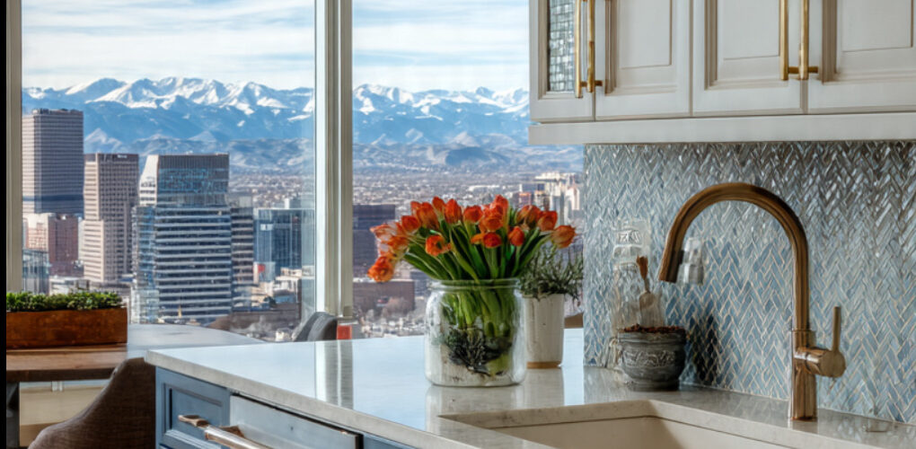 Modern kitchen with blue base cabinets, white uppers, marble countertop, copper faucet, and a city skyline visible through large windows.