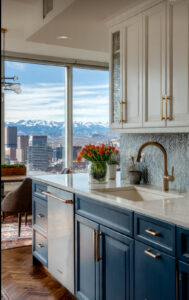 Modern kitchen with blue base cabinets, white uppers, marble countertop, copper faucet, and a city skyline visible through large windows.